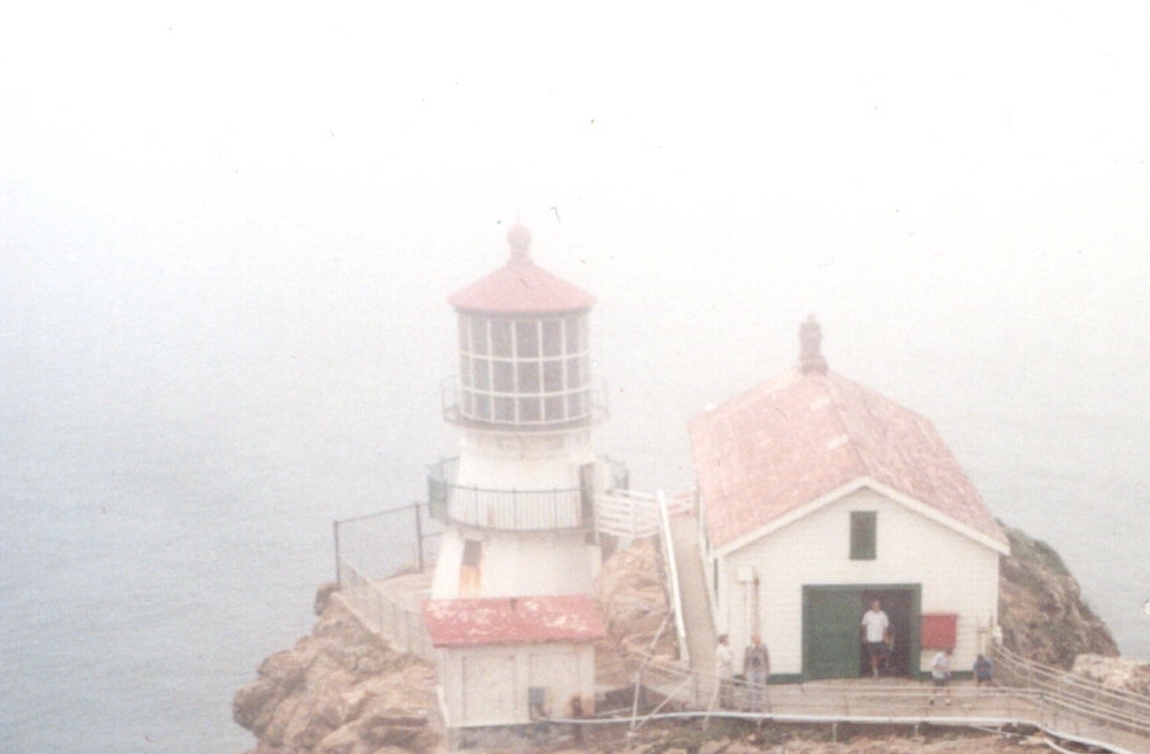 A lighthouse in the fog on a rocky outcrop, with a red roof on the lighthouse and caretaker building, with a person standing in a doorway with a green door open.